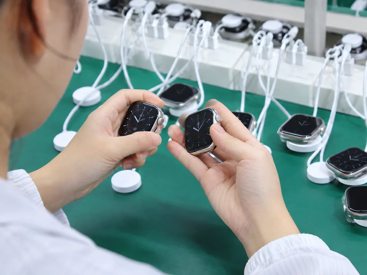 A worker comparing two smartwatches while rows of units undergo charging tests